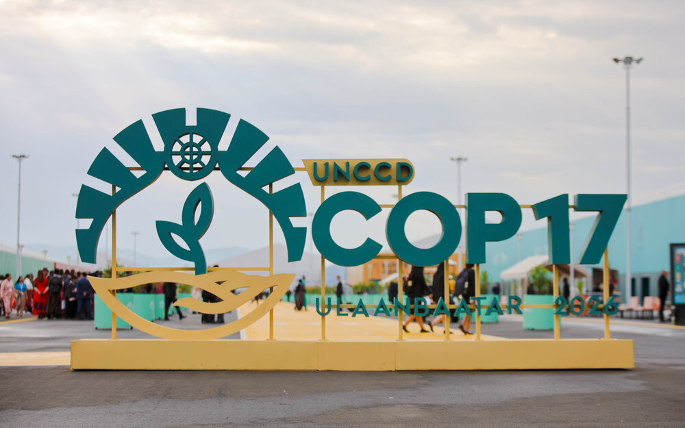Großes türkis-goldenes Schild mit der Aufschrift „UNCCD COP17 ULAANBAATAR 2026“ vor dem Eingang des Veranstaltungsgeländes in Ulaanbaatar. Im Hintergrund sind Besucher, moderne Gebäude und ein bewölkter Himmel zu sehen.